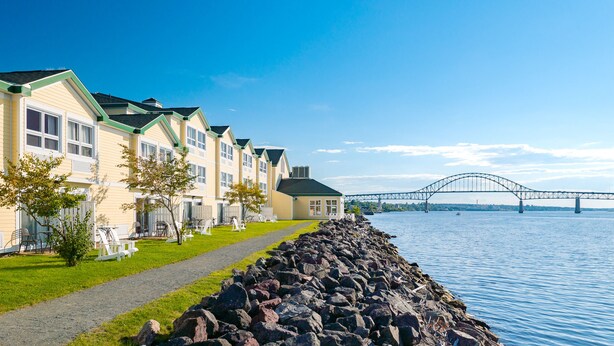 A row of yellow buildings with green roofs by a waterfront, a stone breakwater, and a bridge in the background.