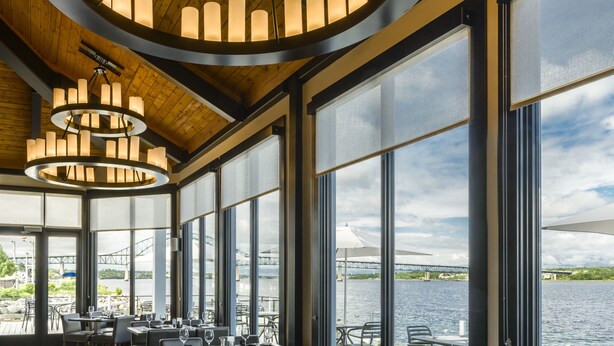 A restaurant with a wooden ceiling, large windows, and a view of the water. Tables are set with chairs and dining ware. - Rodd Miramichi (Miramichi)