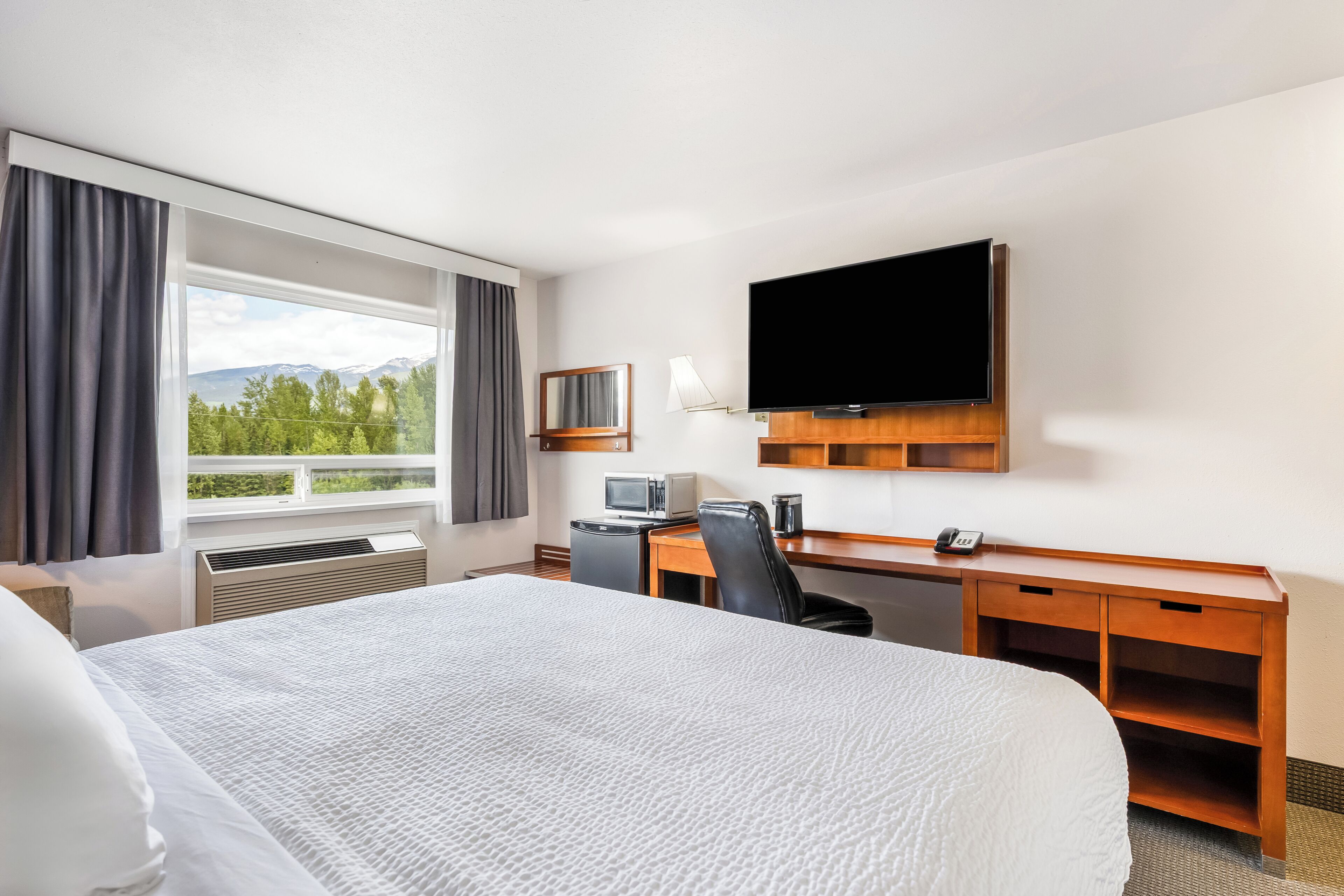 Et hotelværelse med en stor seng, et skrivebord med computer, et fladskærms-tv og et vindue med udsigt over grøn natur og bjerge.