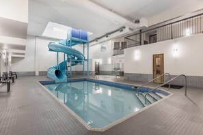 A large indoor swimming pool with a blue slide, a staircase leading to a balcony, and a tiled floor.