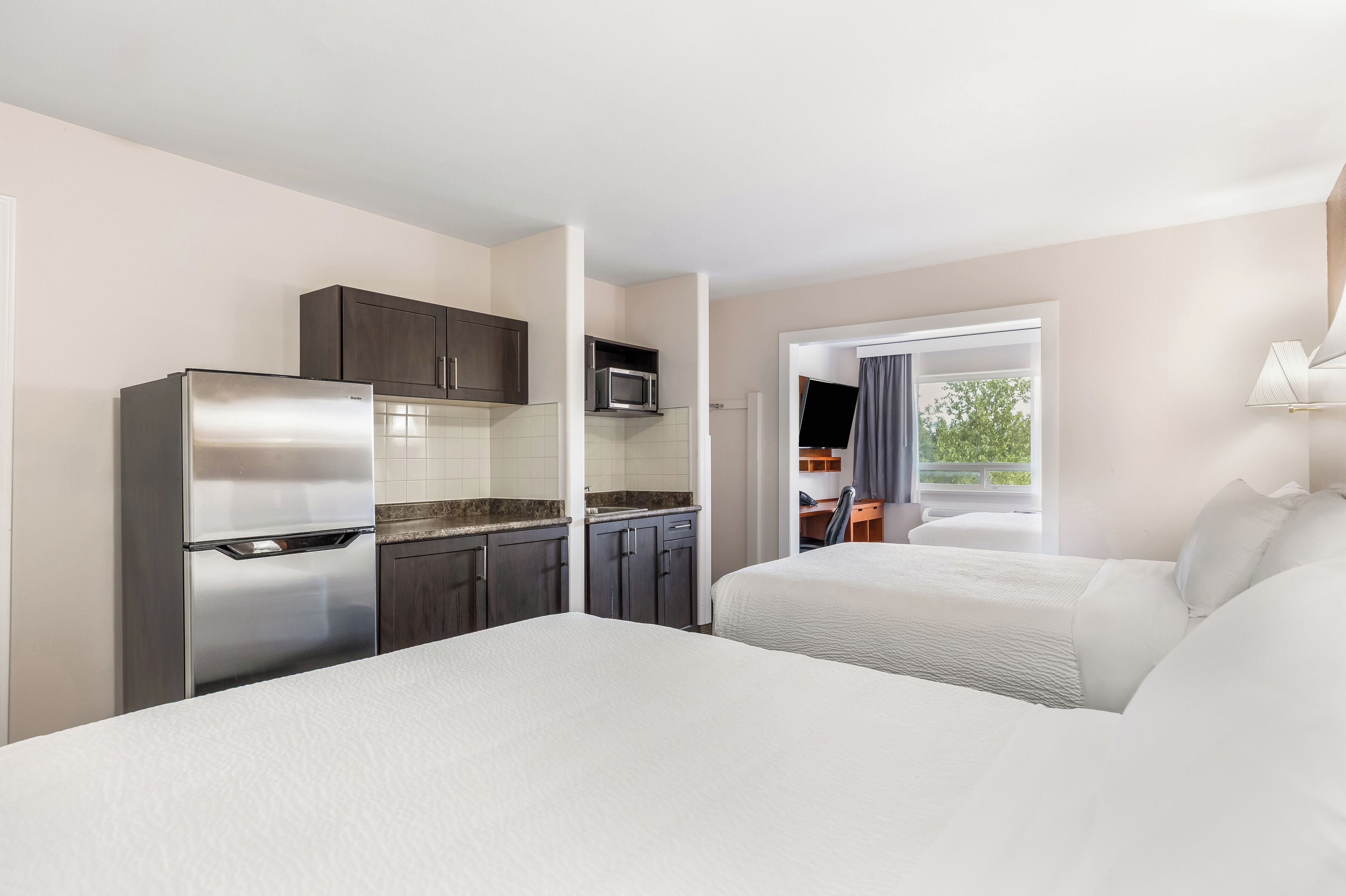 A hotel room with a kitchenette, two beds, a TV, and a window with a view.