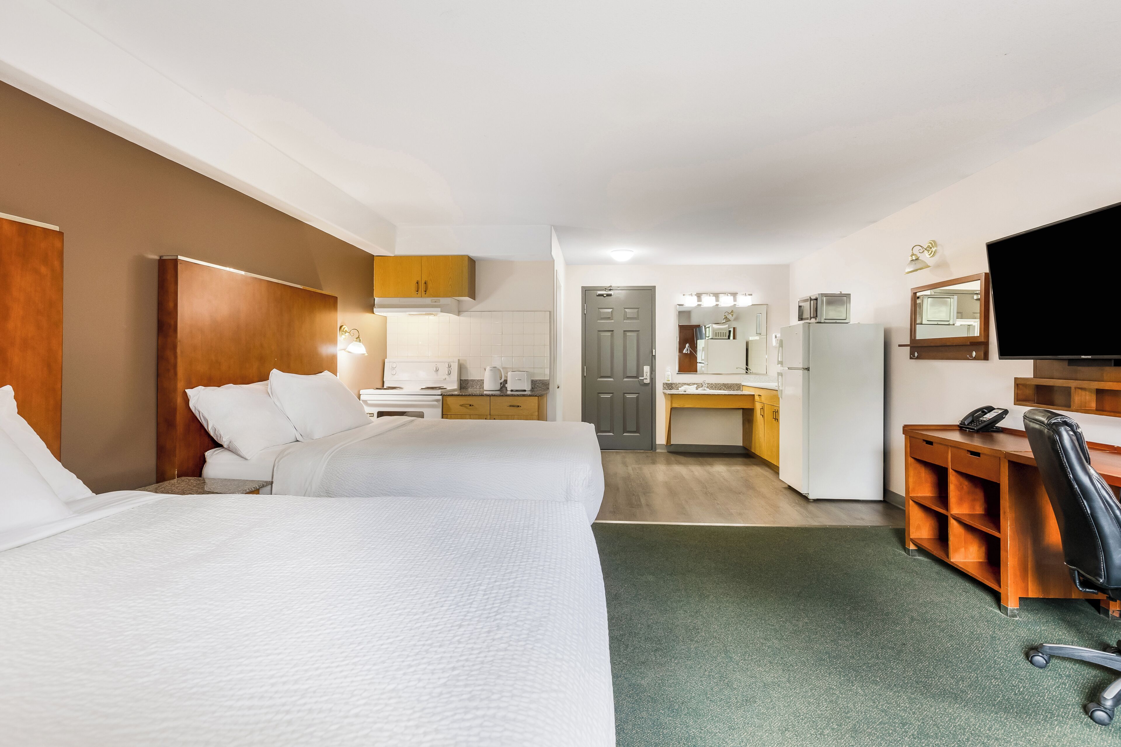 A hotel room with two beds, a desk, a chair, a TV, and a kitchenette.