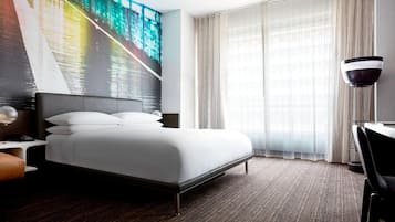 Quarto de hotel com cama, escrivaninha, cadeira, luminária e uma grande janela com cortinas.