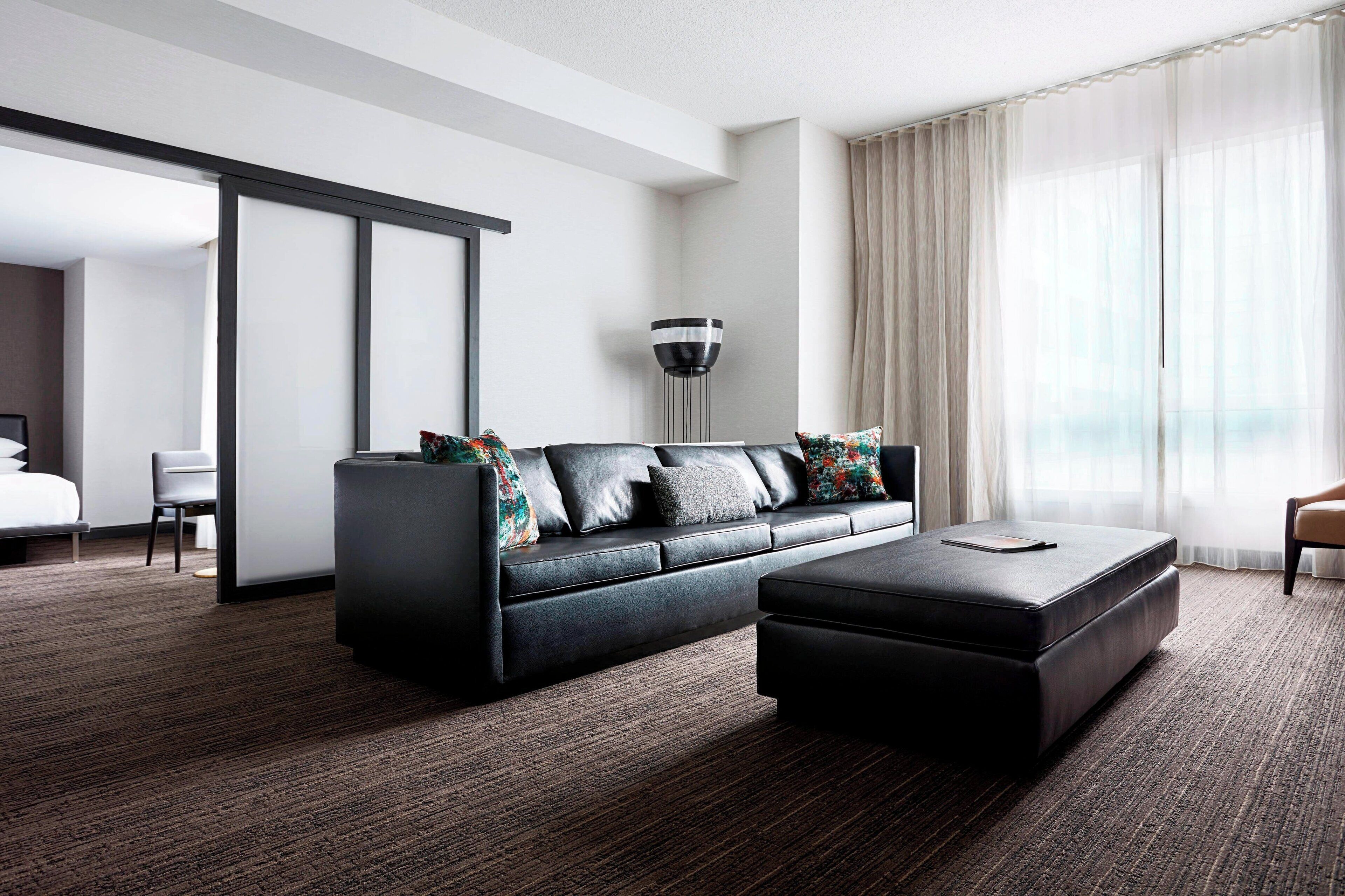 A modern hotel room with a dark leather sofa, a matching ottoman, a large floor lamp, and a view of the city through sheer curtains.