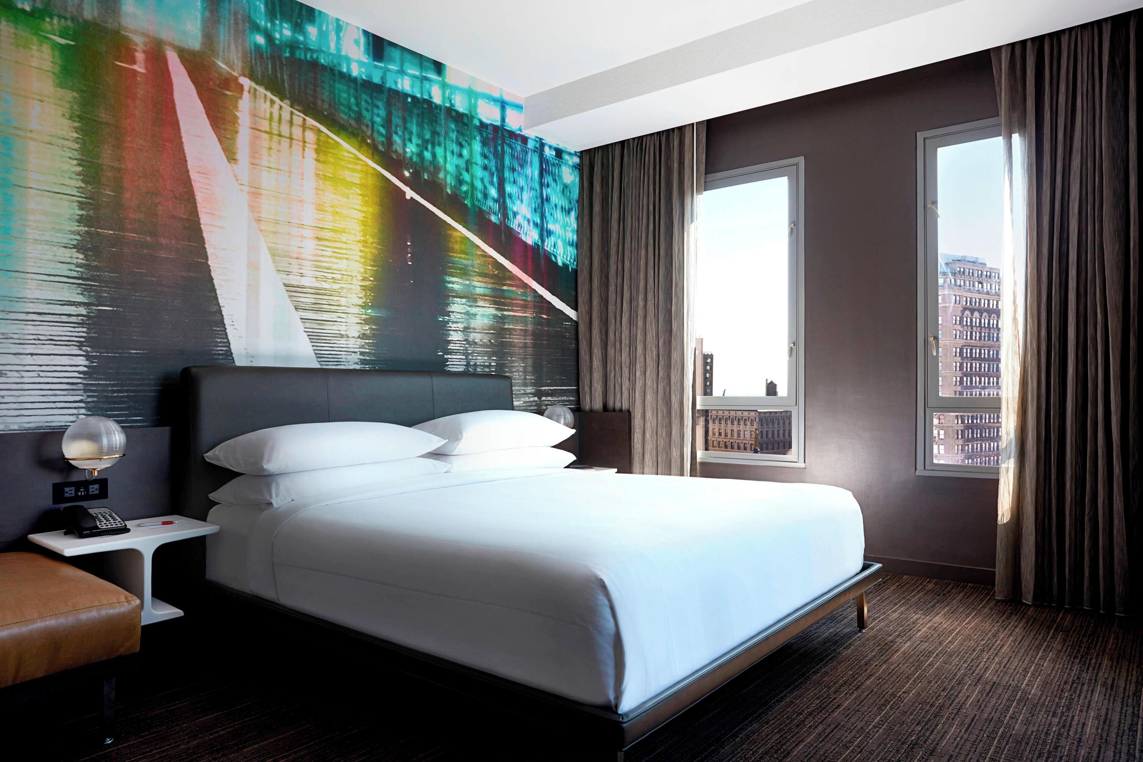 A hotel room with a large bed, a nightstand, a telephone, a window with curtains, and a cityscape view.