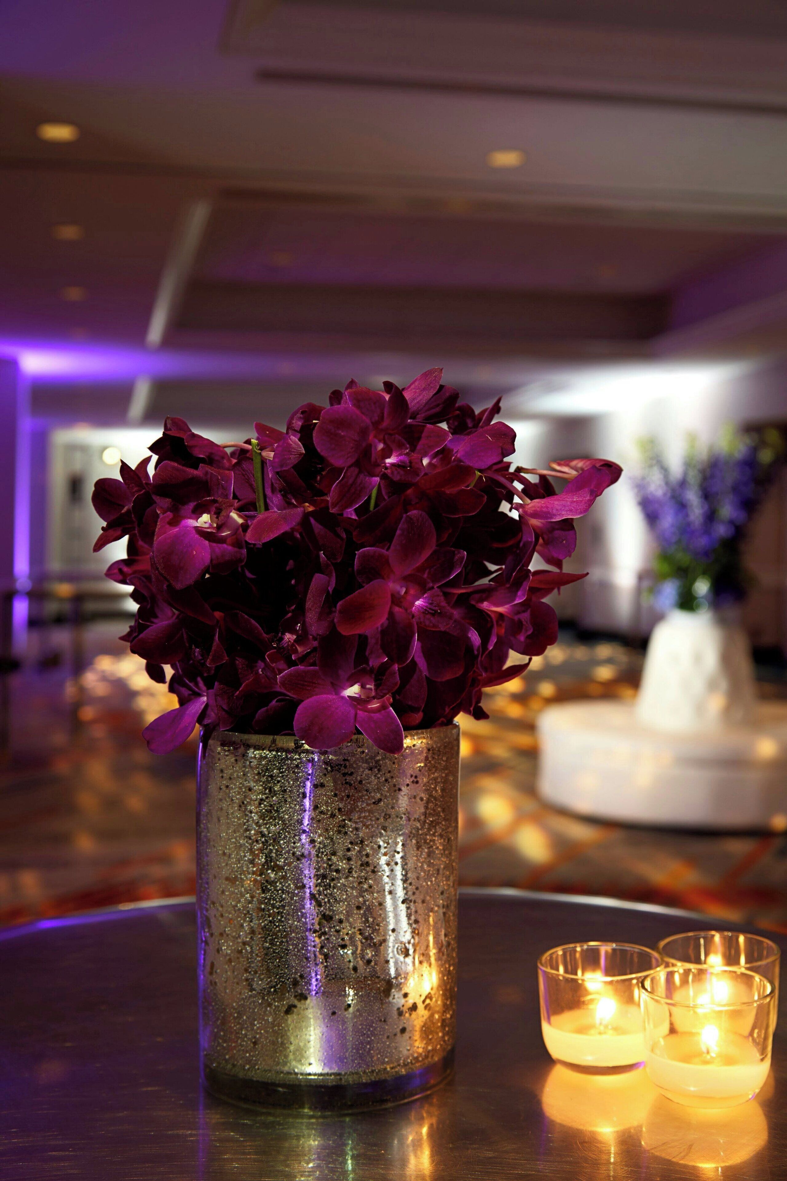 Un florero metálico con flores púrpuras sobre una mesa, junto a dos velas encendidas.