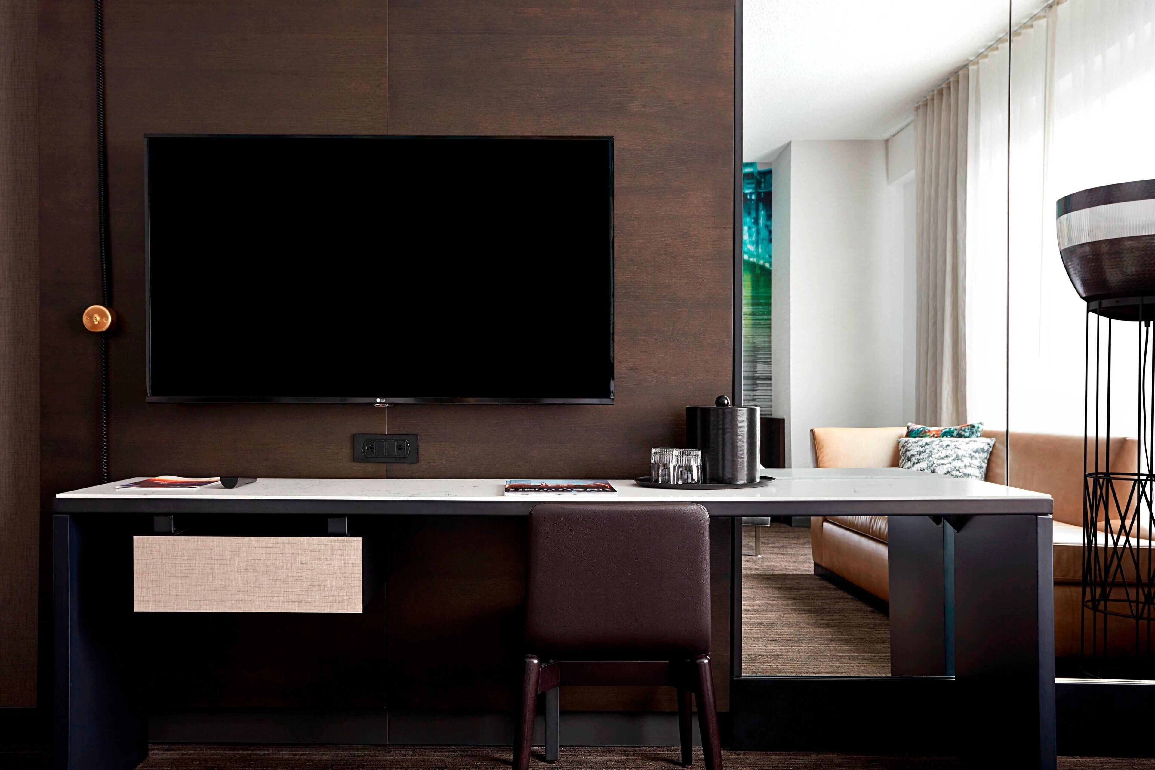 A modern hotel room with a large flat-screen TV mounted on the wall, a dark wooden desk with a chair, a sofa, and a floor lamp.