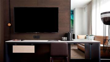 A modern hotel room with a large flat-screen TV mounted on the wall, a dark wooden desk with a chair, a sofa, and a floor lamp.