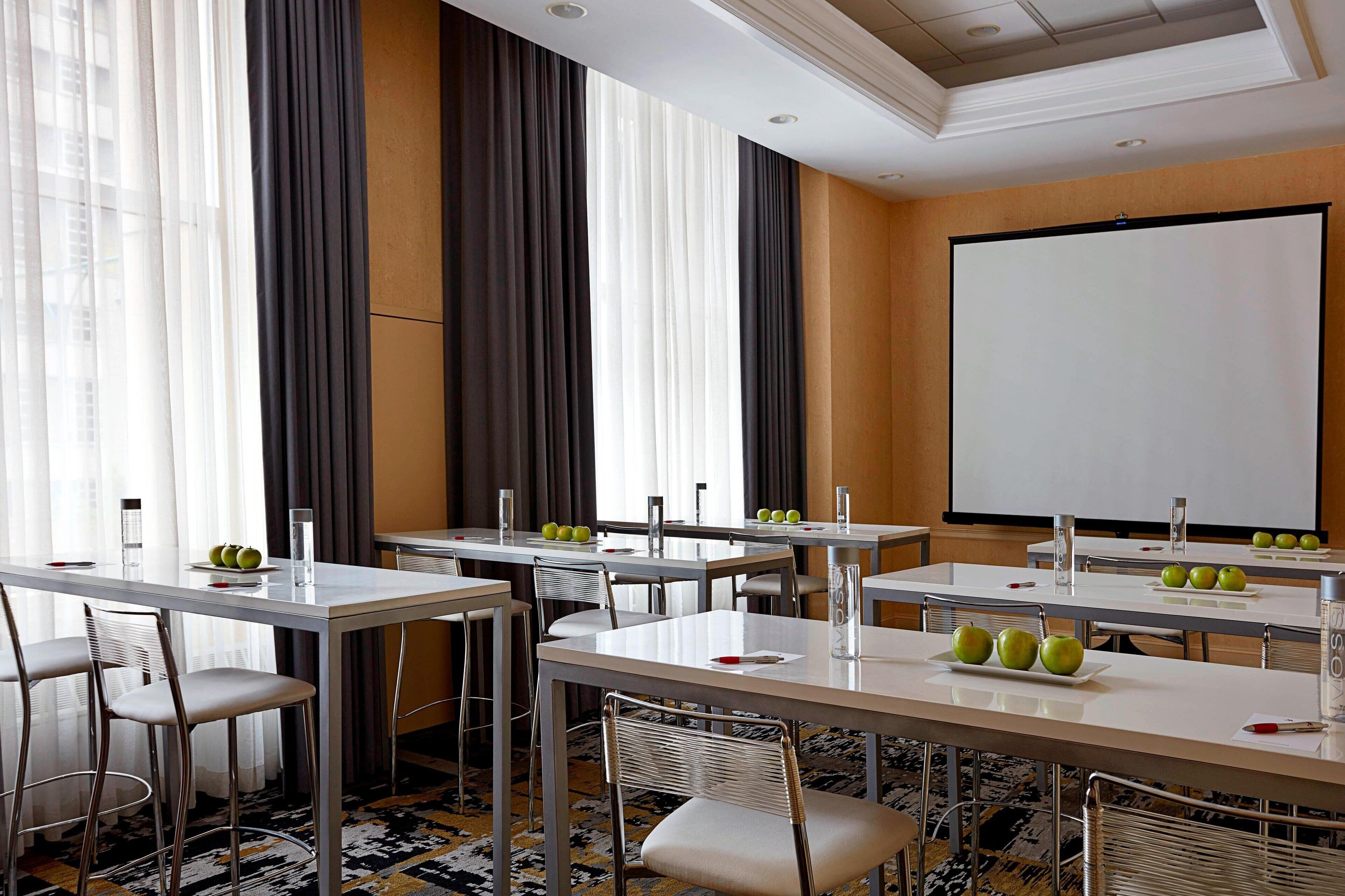 Un salón de conferencias con mesas, sillas, una pantalla de proyector y manzanas sobre las mesas.