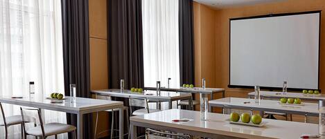 Un salón de conferencias con mesas, sillas, una pantalla de proyector y manzanas sobre las mesas.