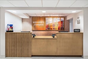 A hotel reception desk with a 'Welcome to Hampton' sign, a digital kiosk, and a 'Hilton Honors' sign. - Hampton Inn White River Junction (White River Junction)