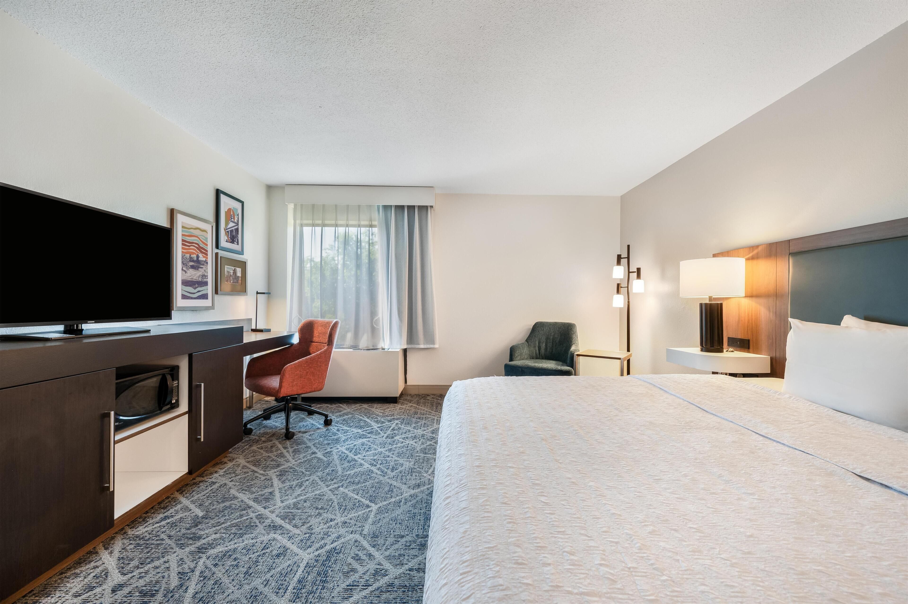A hotel room with a bed, a desk with a flat-screen TV, a chair, a lamp, and a window with curtains.