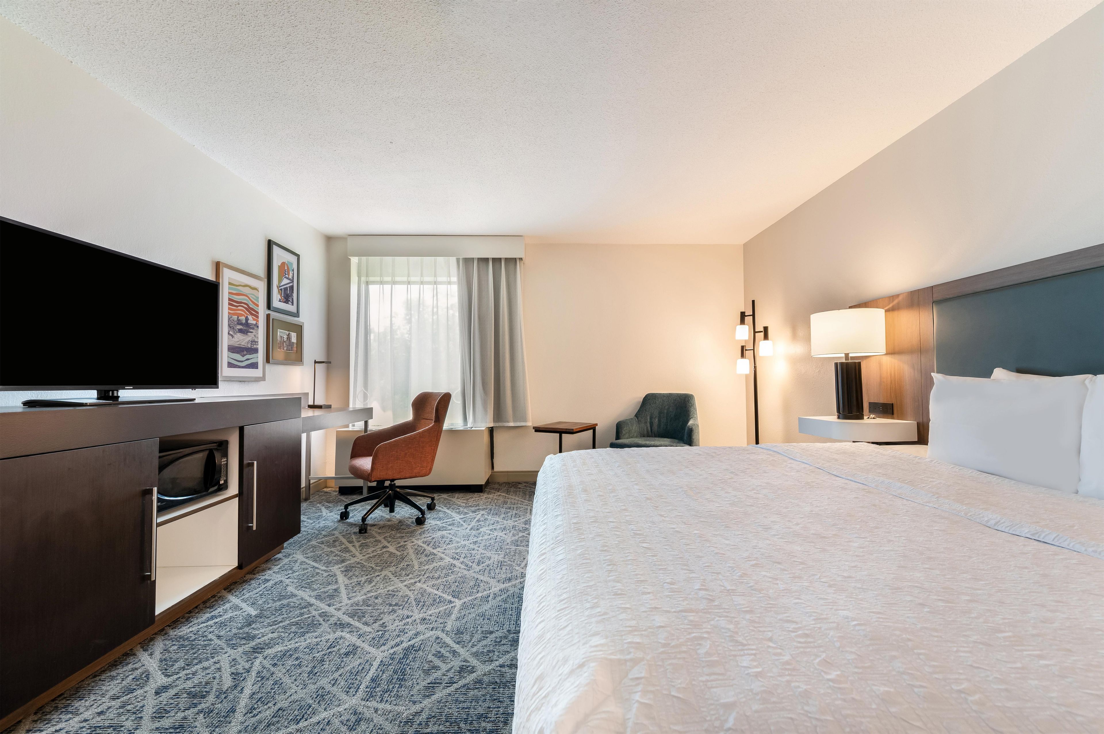 Habitación de hotel con cama, escritorio, silla, televisión y ventana con cortinas.