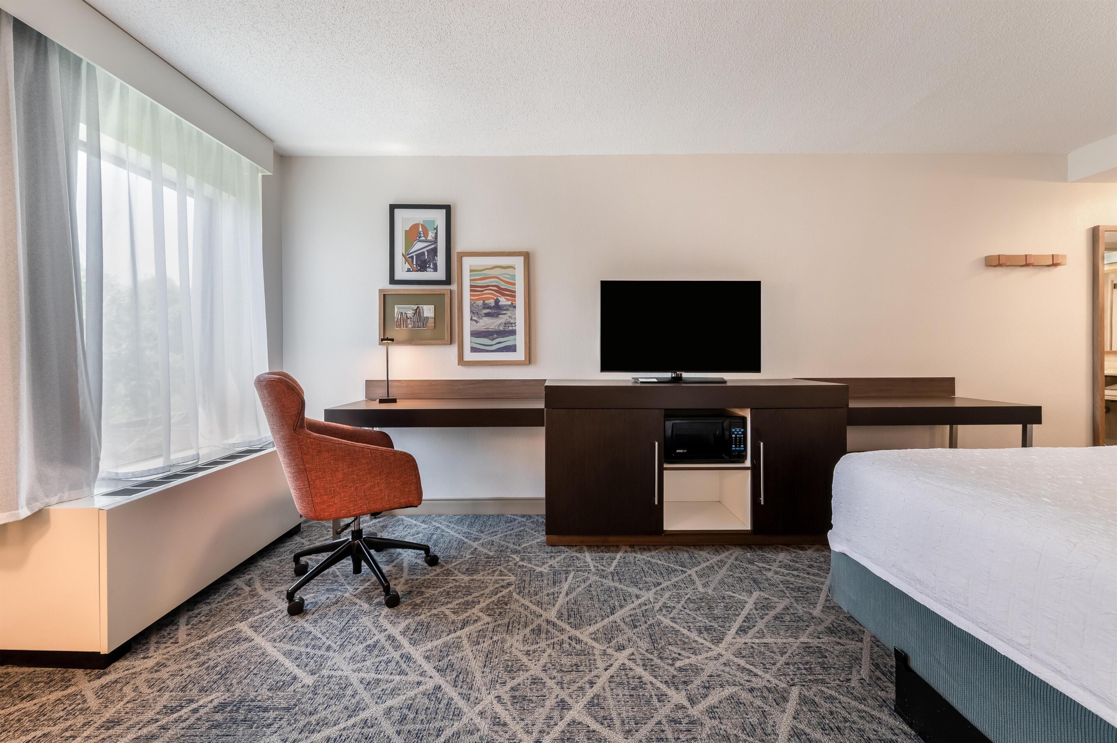 Habitación de hotel con cama, escritorio con televisor de pantalla plana, una silla y ventana con cortinas traslúcidas.