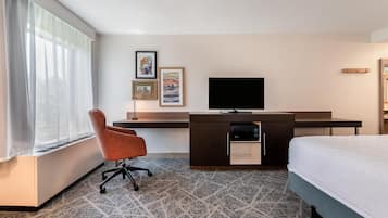 Habitación de hotel con cama, escritorio con televisor de pantalla plana, una silla y ventana con cortinas traslúcidas.