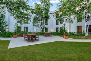 Un patio exterior con mesa y sillas, rodeado de follaje y una fachada de edificio moderna.