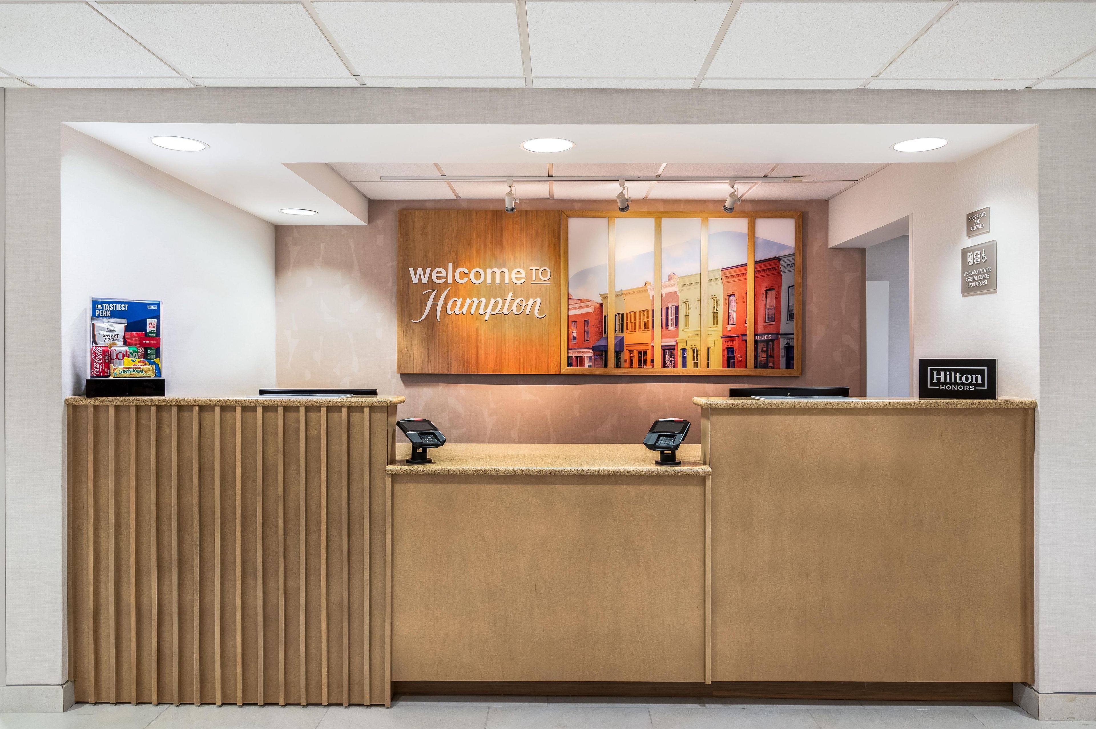 A hotel reception desk with a 'Welcome to Hampton' sign, a digital kiosk, and a 'Hilton Honors' sign.
