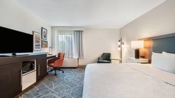 A hotel room with a bed, a desk with a flat-screen TV, a chair, a lamp, and a window with curtains.
