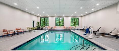 An indoor swimming pool with a tiled floor, surrounded by chairs and tables, and large windows with a view of trees.