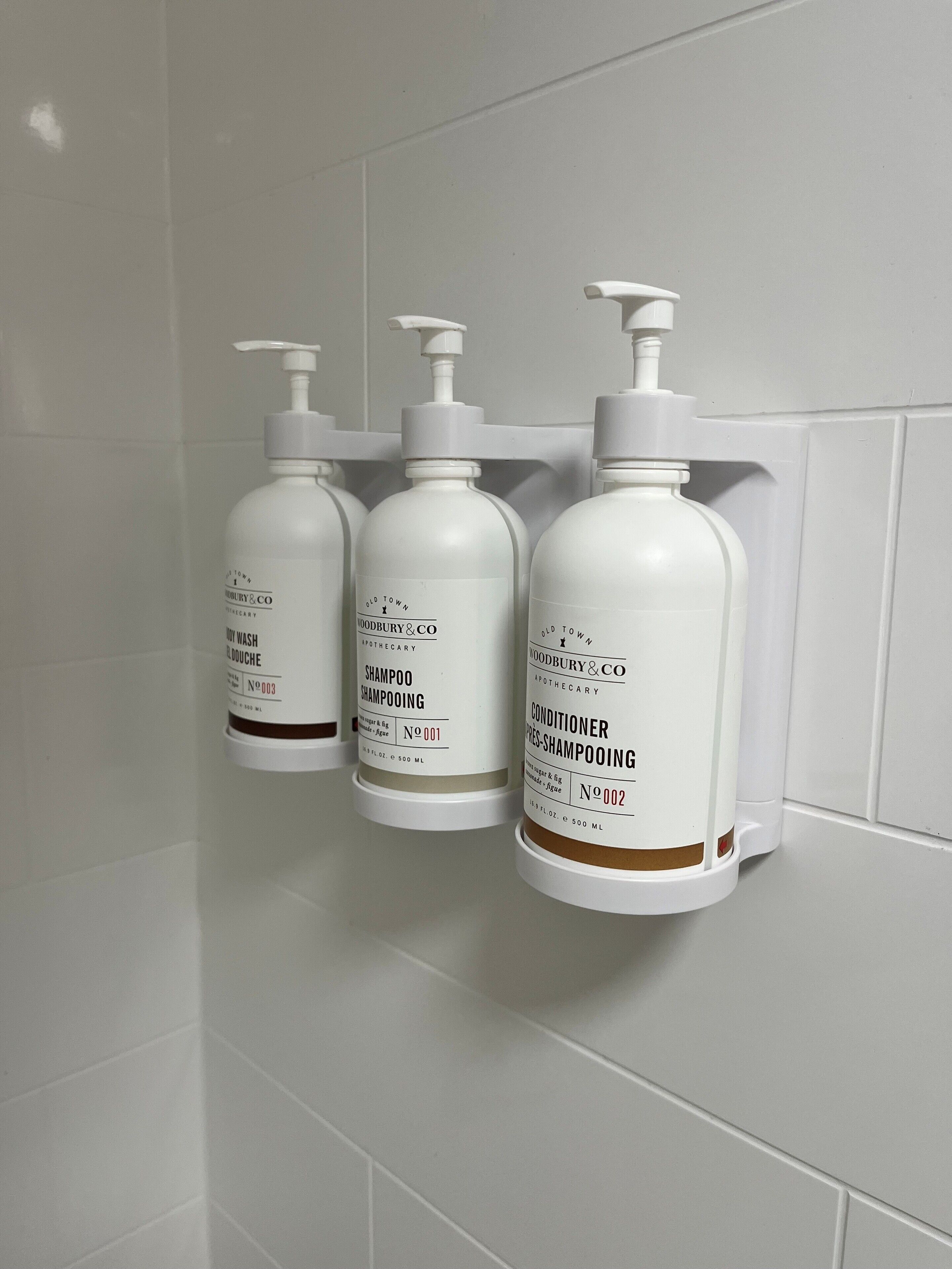 Tres botellas blancas de productos Woodbury & Co. sobre una pared de azulejos blancos.