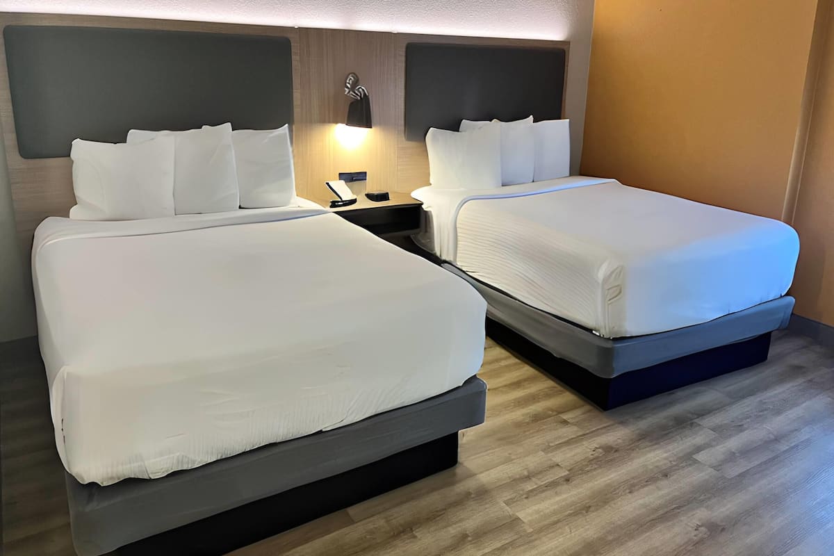 A hotel room with two beds, each with white bedding and headboards. There is a nightstand with a lamp between the beds.