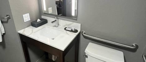 A bathroom with a white sink, mirror, and toilet. There is a handrail on the wall and a towel rack.