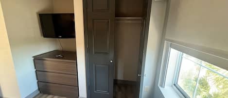 A compact room with a dark door, a small dresser, a television mounted on the wall, and a window with a view of greenery outside.