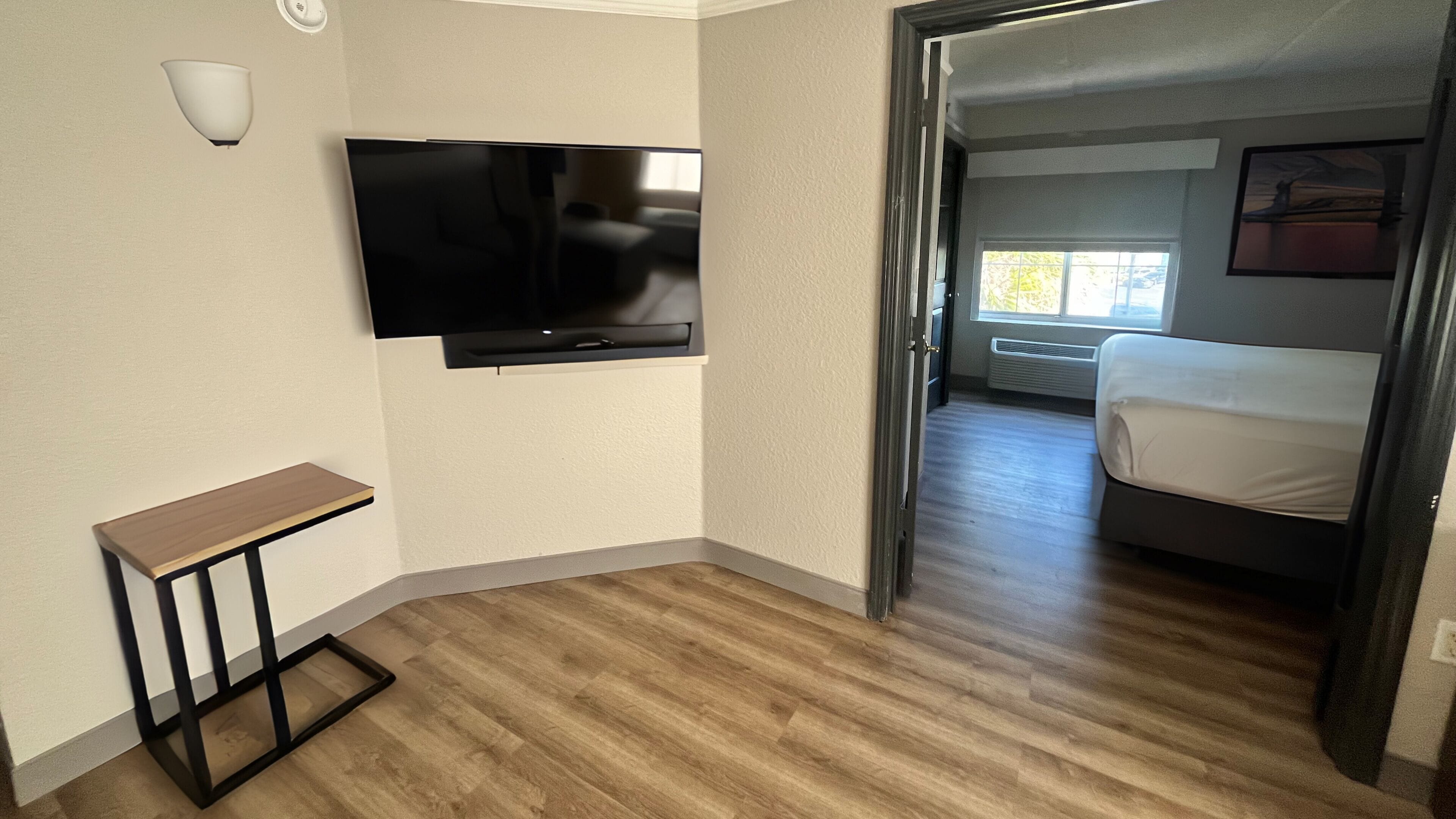 A hotel room with a flat-screen TV mounted on the wall, a small wooden table, and a bed with a white mattress.