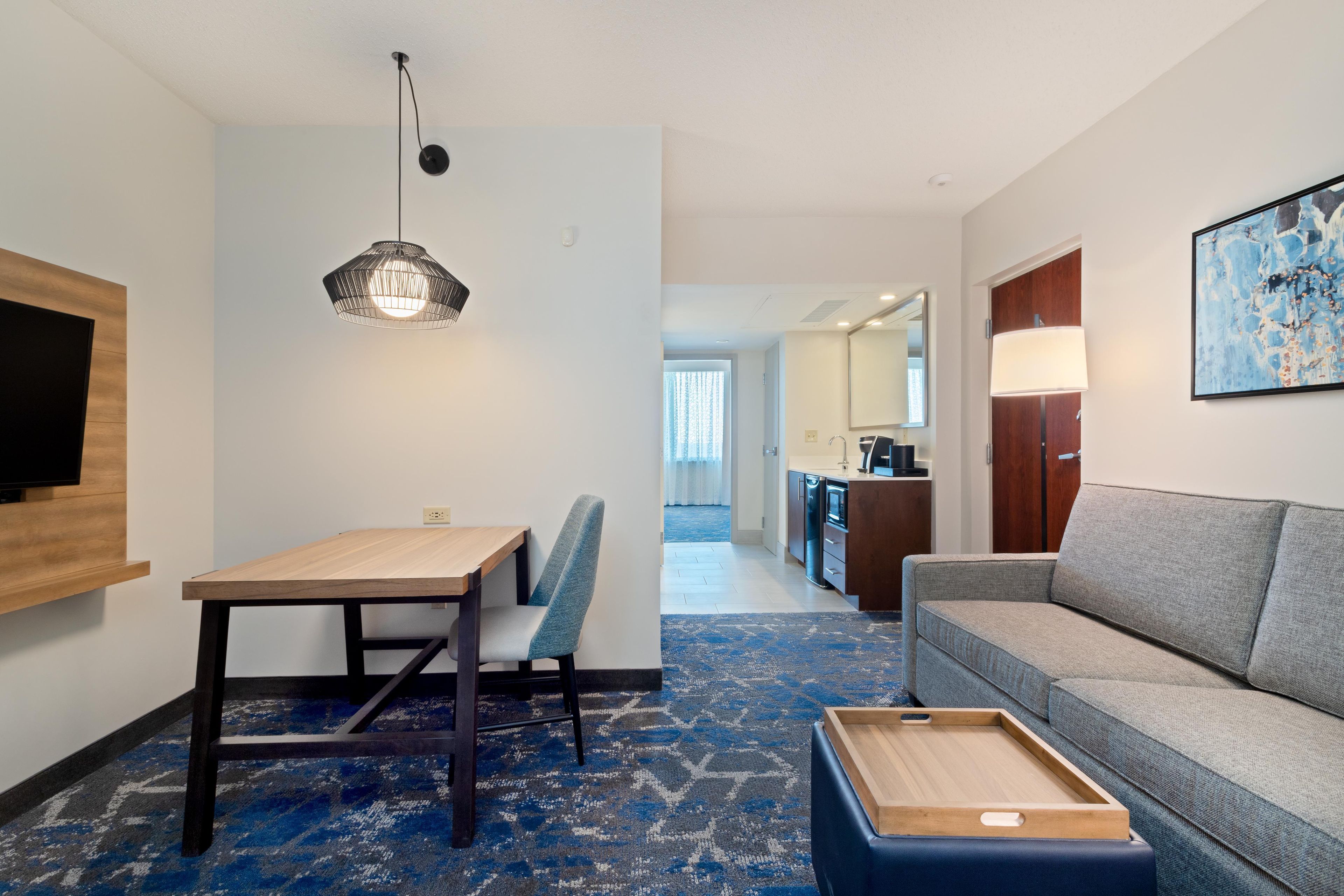 Habitación de hotel con una mesa de comedor, un sofá, un televisor montado en la pared y una cocina visible a través de una puerta abierta.