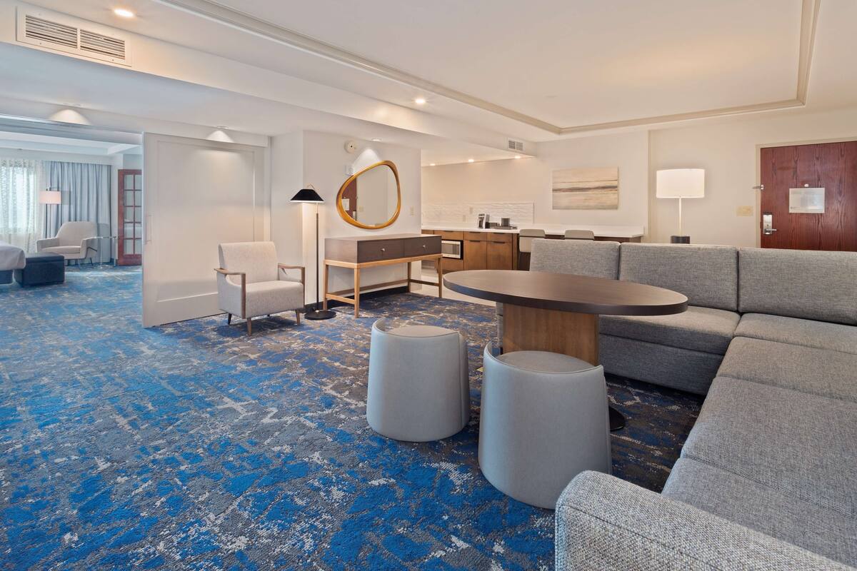 Ein Hotelzimmer mit blauem Teppich, einem grauen Sofa, einem runden Tisch und einem Holzschrank.