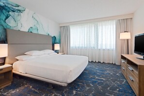 Ein Hotelzimmer mit einem großen Bett, einem Flachbildfernseher, einem Nachttisch, einer Lampe und einem Fenster mit Vorhängen.