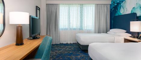Habitación de hotel con escritorio, dos camas, televisor y un ventanal grande con cortinas.
