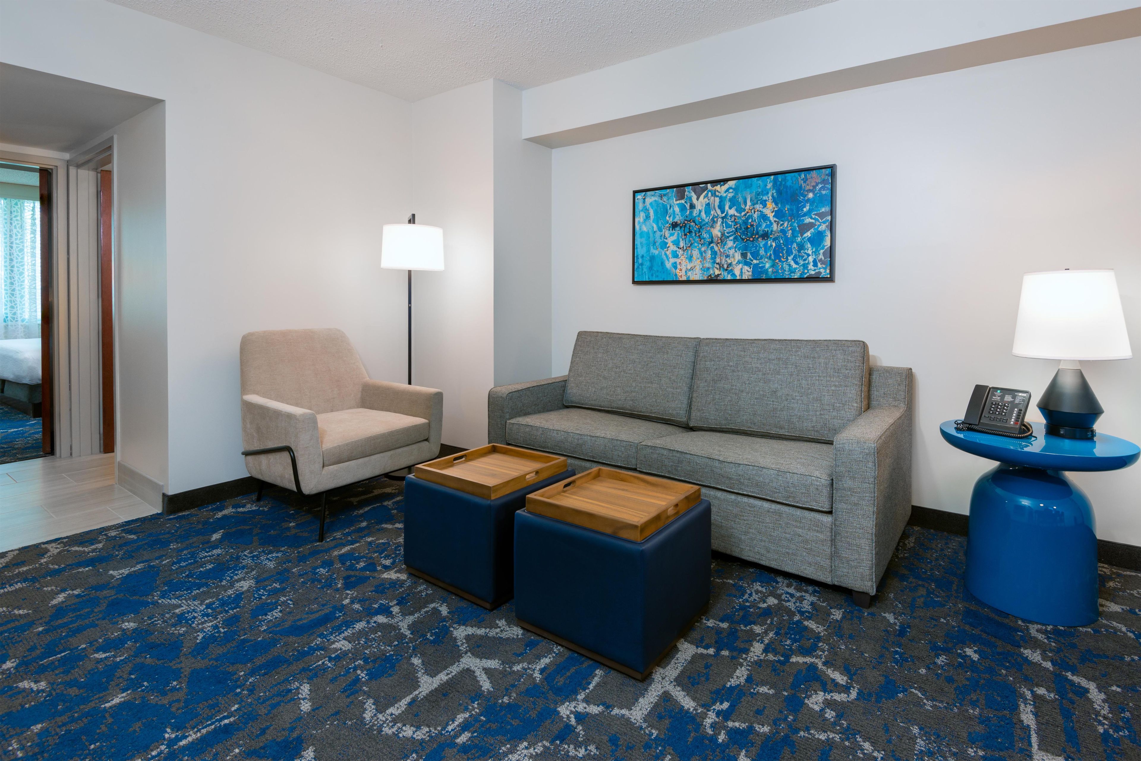 Habitación de hotel con sofá, sillón, dos otomanas, un cuadro, una lámpara y un teléfono sobre una mesa azul.