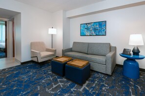 Habitación de hotel con sofá, sillón, dos otomanas, un cuadro, una lámpara y un teléfono sobre una mesa azul.