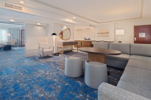 Ein Hotelzimmer mit blauem, gemustertem Teppich, einem Sofa, einem runden Tisch, einem Schreibtisch und einem Spiegel.
