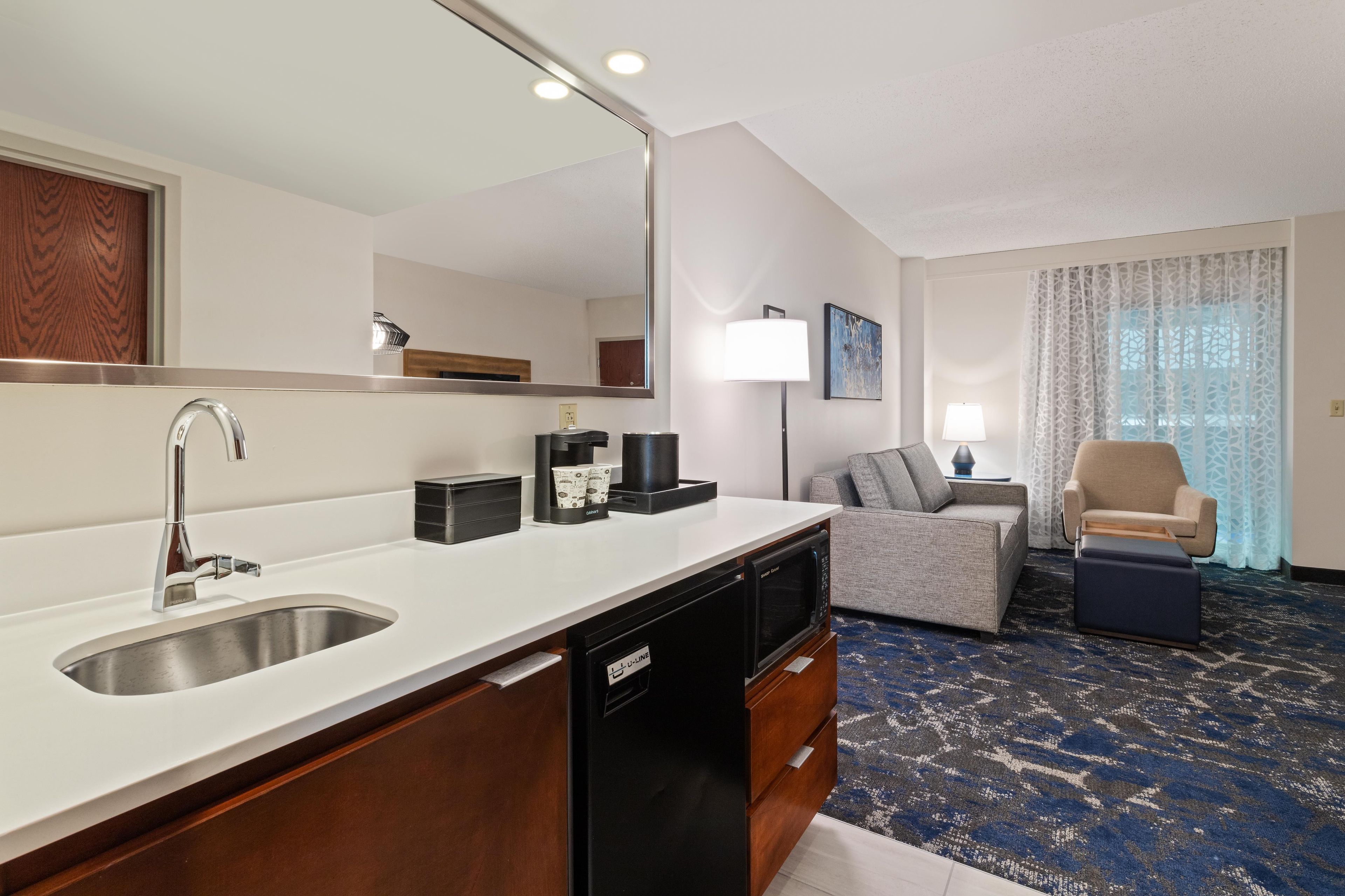 Habitación de hotel moderna con cocina, zona de estar con sofá y sillones, y un espejo grande.