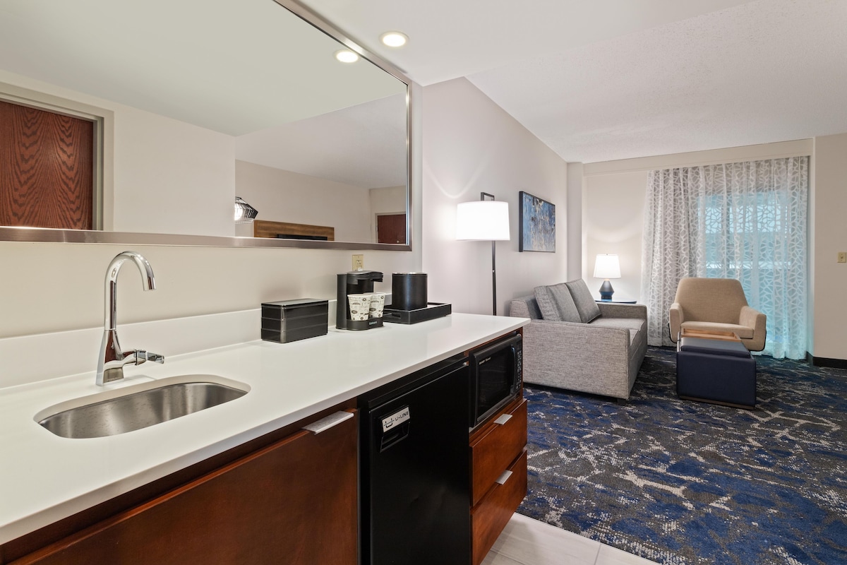 Une chambre d’hôtel moderne dotée d’un coin cuisine, d’un espace salon avec un canapé et des fauteuils, et d’un grand miroir.