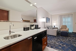 Een moderne hotelkamer met een keukenhoekje, een zithoek met een bank en fauteuils, en een grote spiegel.