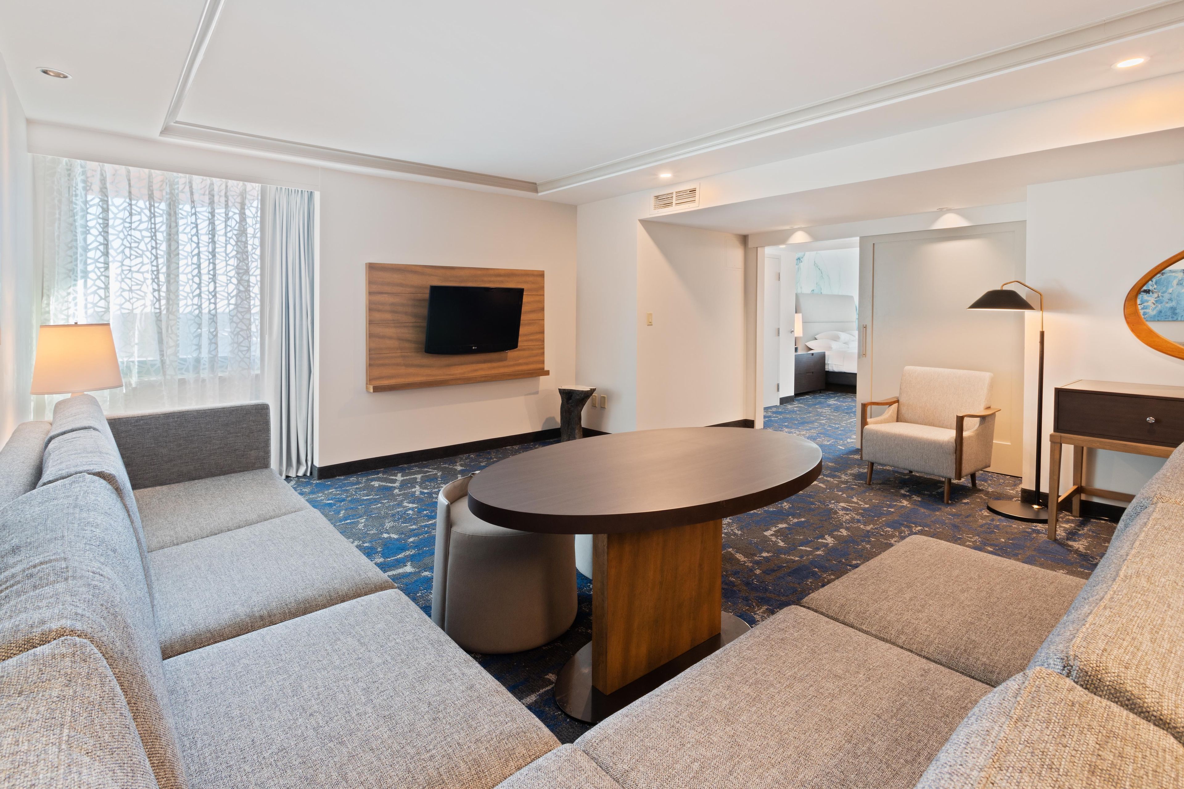 Habitación de hotel moderna con sofá, una mesa redonda, televisor de pantalla plana montado sobre un panel de madera, lámpara y vista al pasillo.