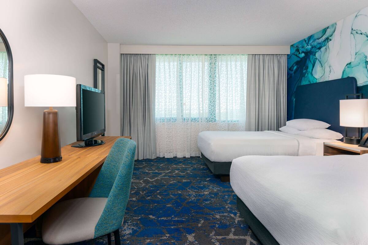Ein Hotelzimmer mit zwei Betten, einem Schreibtisch mit Computer, einem Stuhl und einem großen Fenster mit Vorhängen.