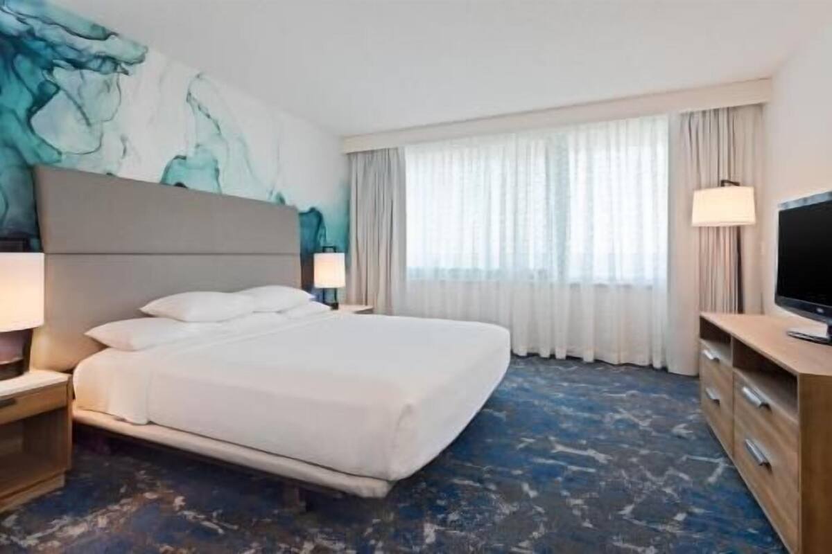 Ein Hotelzimmer mit Bett, Fernseher, Schreibtisch und einem großen Fenster mit Vorhängen.