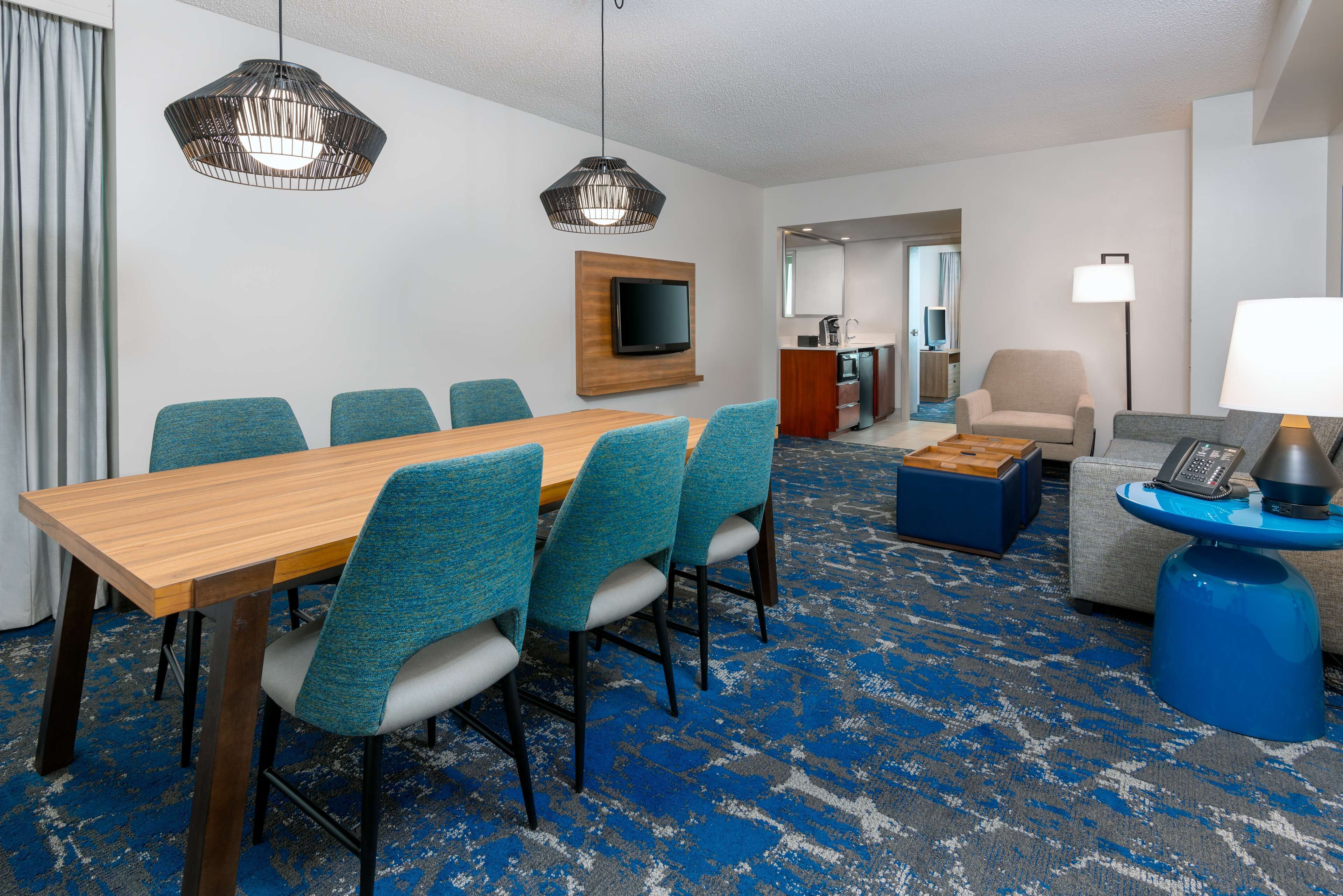 A hotel room with a wooden dining table, blue upholstered chairs, a blue patterned carpet, a television mounted on the wall, a sofa, a coffee table, and a lamp.