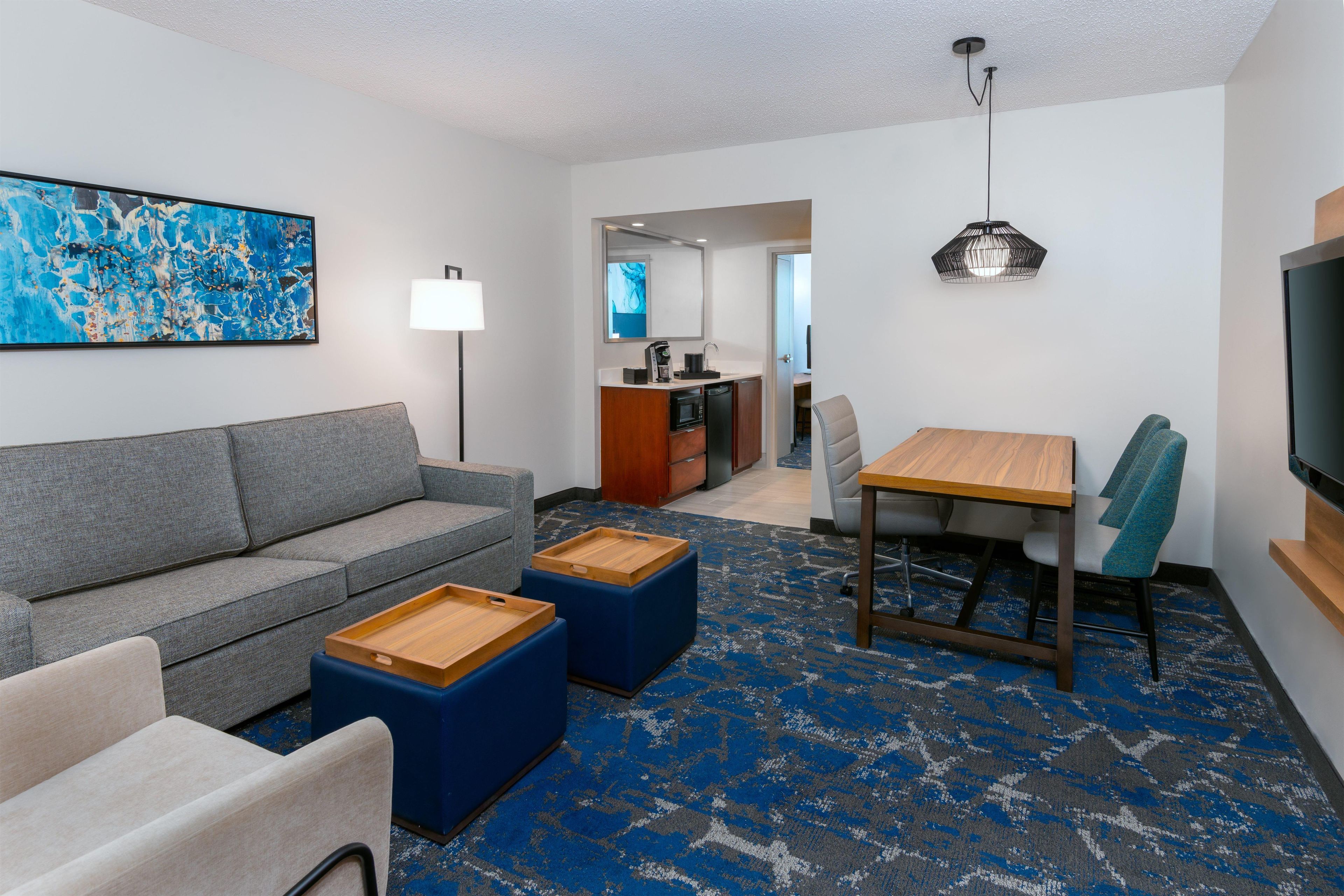 Ein modernes Hotelzimmer mit einem grauen Sofa, einem Holztisch und einem blau gemusterten Teppich.