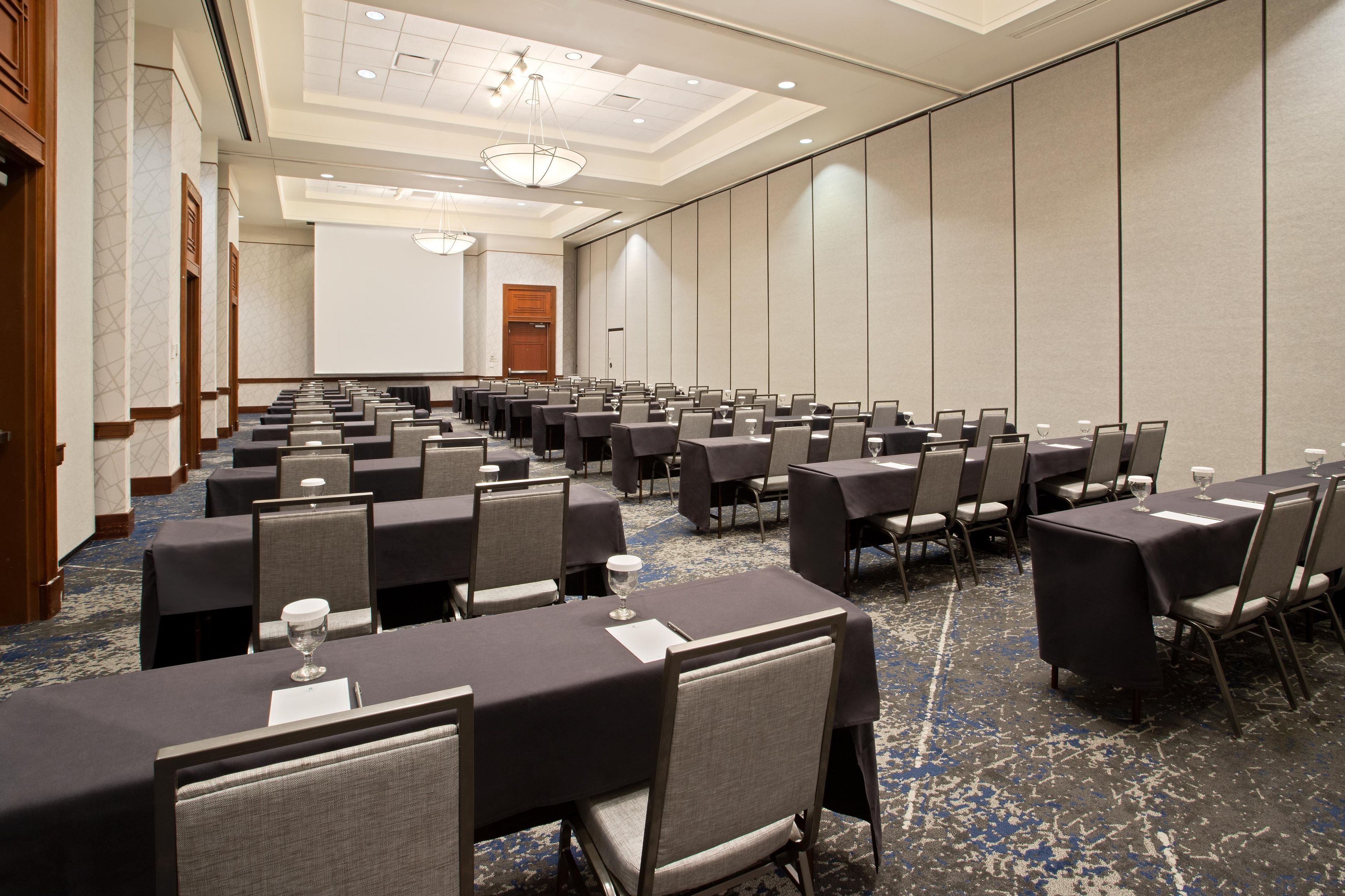 A conference room with rows of tables and chairs, a projector screen, and a large door.