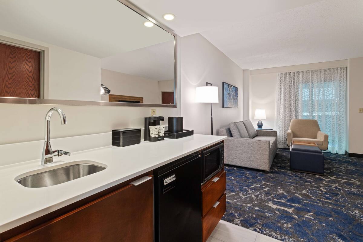 Ein modernes Hotelzimmer mit Küchenbereich, einer Couch, einem Sessel und einem großen Fenster mit Vorhängen.