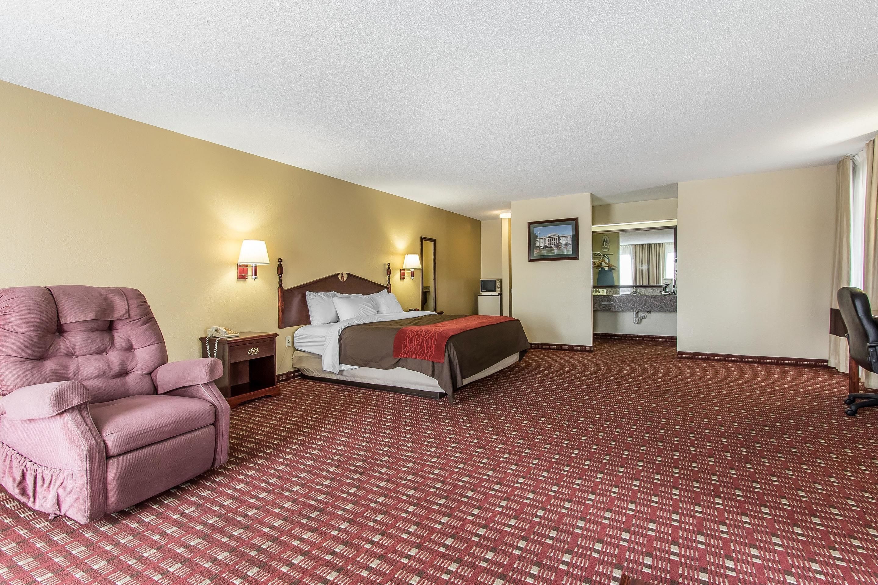 A hotel room with a large bed, a pink armchair, a desk, and a bathroom visible through an open door.