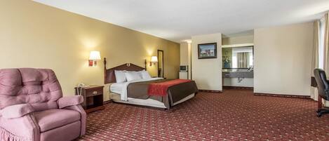 A hotel room with a large bed, a pink armchair, a desk, and a bathroom visible through an open door.