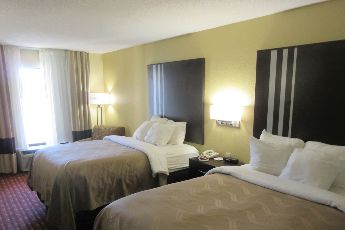 A hotel room with two beds, a bedside table with a lamp, a telephone, and a wall-mounted light fixture.