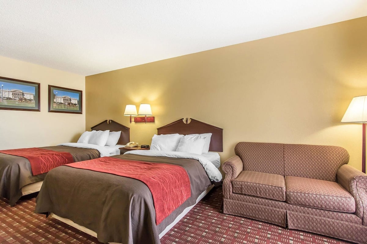 A hotel room with two beds, a sofa, a lamp, and two wall-mounted lamps.