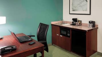 A conference room with a wooden table, a laptop, a projector, a black office chair, a kitchenette with a microwave, a sink, and a framed picture on the wall.